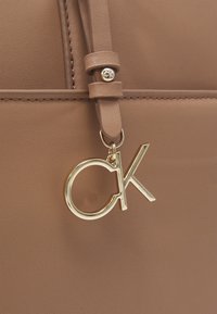 Braune Kunstleder-Handtasche mit glatter Textur, ausgestattet mit einem goldenen "CK"-Logo-Anhänger und einer Hardware-Akzentuierung am Riemen.