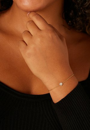 Bracelet chaîne en or avec un ornement central semblable à un diamant, porté au poignet sur un fond de tissu côtelé sombre.