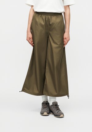 Άτομο φοράει ελαφρώς φαρδιά παντελόνια σε ελιά, λευκές κάλτσες, γκρίζα αθλητικά παπούτσια και λευκό κοντομάνικο τοπ σε απλό φόντο.