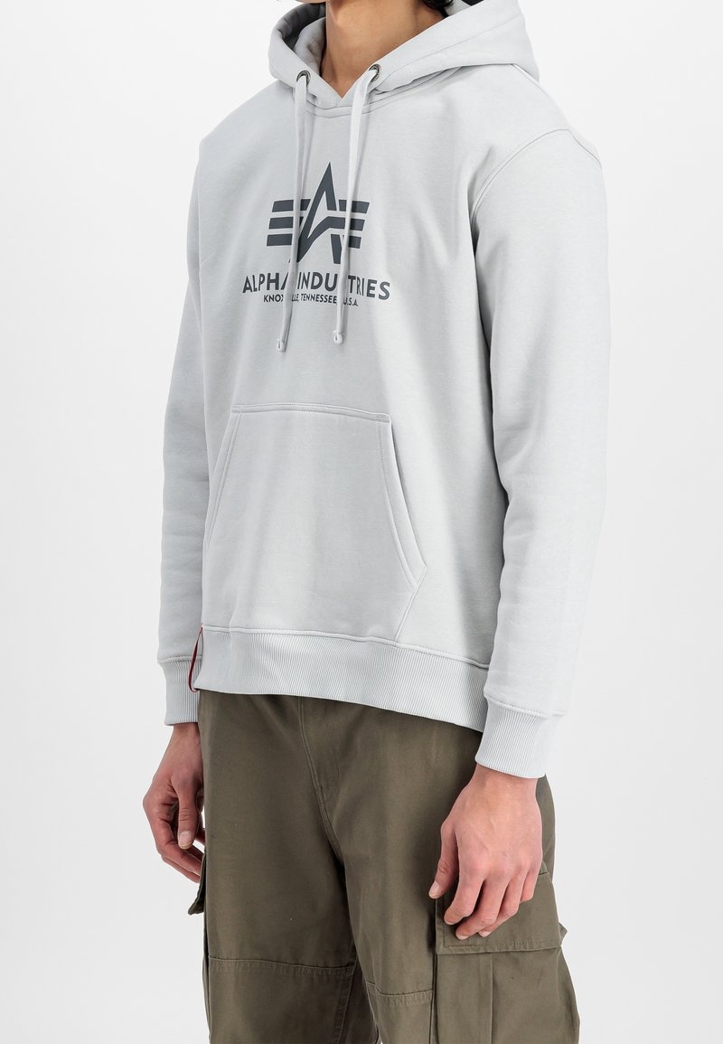 Homme portant un sweat à capuche Alpha Industries gris clair avec une poche avant et un pantalon cargo vert olive, debout devant un fond blanc.