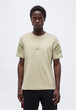 Άνδρας που φοράει μπεζ κοντομάνικη μπλούζα με τρεις πράσινες ρίγες σε κάθε μανίκι και ένα μικρό πράσινο λογότυπο στο στήθος, στέκεται μπροστά σε λευκό φόντο.