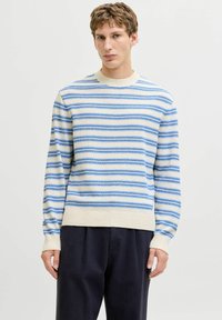 Homme portant un pull rayé crème et bleu avec un pantalon noir, debout devant un fond clair uni avec une expression neutre.