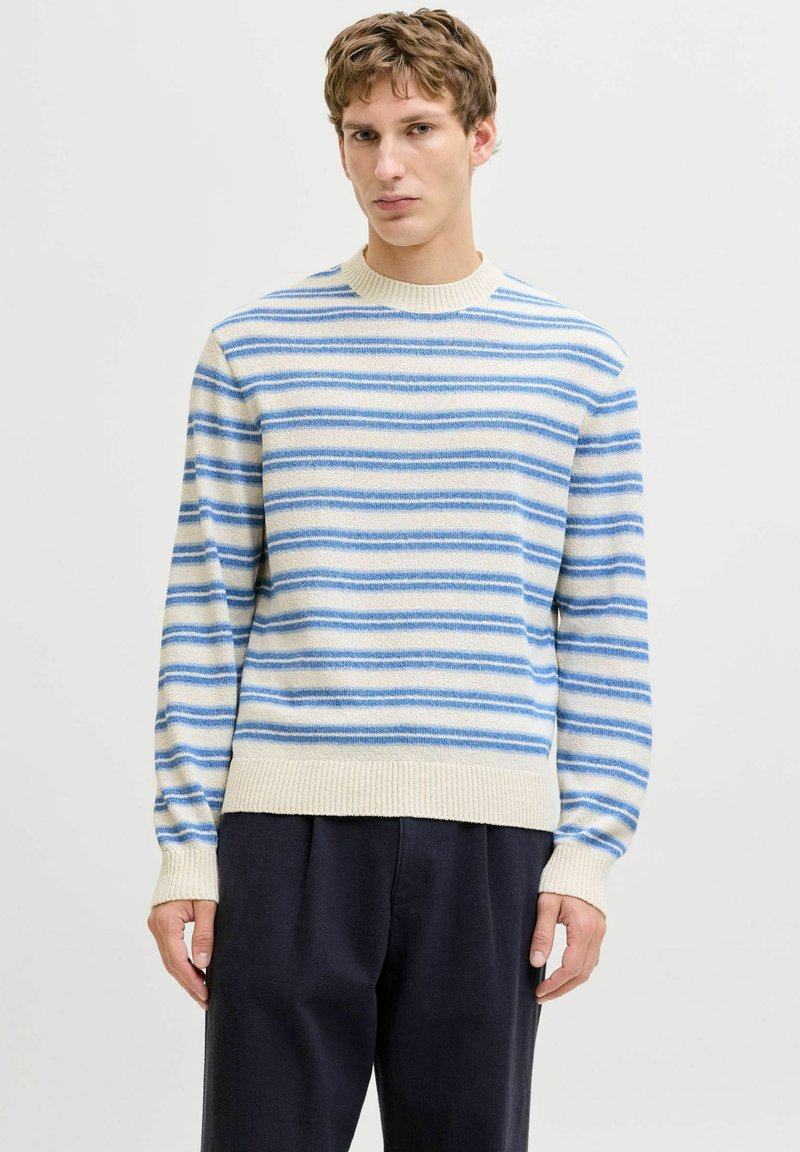 Homme portant un pull rayé crème et bleu avec un pantalon noir, debout devant un fond clair uni avec une expression neutre.