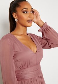 Fialové šaty s hlubokým výstřihem do V, dlouhými průsvitnými rukávy a nařaseným pasem. Vyrobeno z měkké, lehké tkaniny a s elastickými manžetami rukávů.