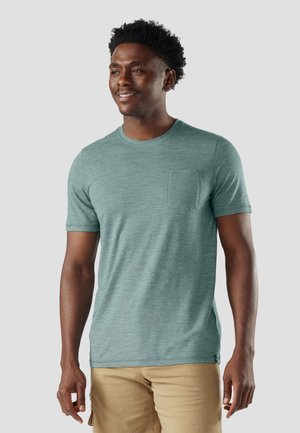 Lächelnder Mann mit kurzen lockigen Haaren, der ein grünes kurzärmeliges T-Shirt mit Tasche und beigen Shorts vor einem einfarbigen Hintergrund trägt.