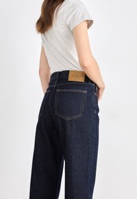 Mörka denimjeans med rak passform, med bakfickor och en bärnstenfärgad läderetikett vid midjan. Grå t-shirt delvis synlig.