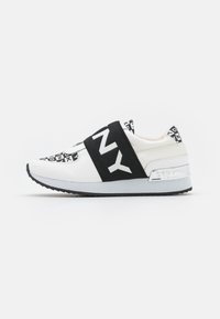Weiße Sportschuh mit schwarzem Riemen, auf dem der Text "DKNY" steht. Glattes Obermaterial, graue Laufsohle und metallischer Absatzakzent.