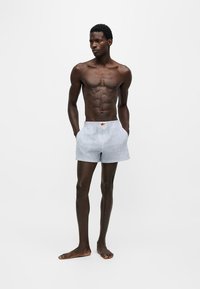 Homme mince et musclé debout pieds nus, portant un short rayé bleu clair et blanc, les mains dans les poches sur fond blanc uni.