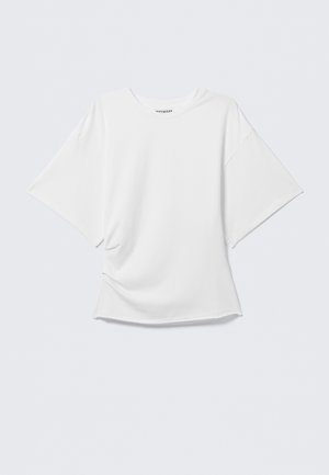 Hvitt oversized T-skjorte med kort erme og rund hals, lett krøllet foran, vist mot en ensfarget lys grå bakgrunn.
