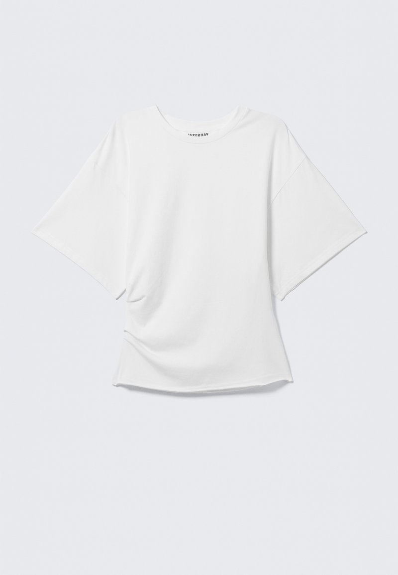 T-shirt blanc oversize à manches courtes avec col rond, devant légèrement froissé, présenté sur un fond gris clair uni.