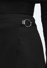 Gros plan sur un pantalon noir taille haute avec une boucle circulaire en métal argenté à la ceinture, porté avec un haut noir.