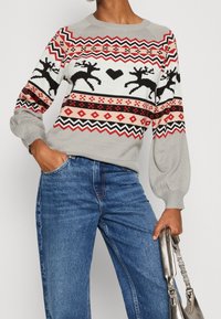 Szary dziany sweter z wzorem świątecznym przedstawiającym renifery i serca w kolorze czerwonym, czarnym i białym. Luźny krój z bufiastymi rękawami. Niebieskie dżinsy.