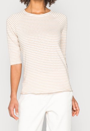 Vrouw draagt een beige-wit met horizontale strepen halfmouwtop en witte broek, staand tegen een effen lichte achtergrond.