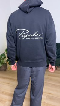 Black hoodie with white "Pegador" text on the back, features a kangaroo pocket and ribbed cuffs, paired with dark pants.