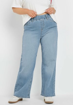 Frau zieht am elastischen Bund einer hellblauen Jeans mit weitem Bein, trägt weiße Sneakers und ein weißes, hineingestecktes T-Shirt.