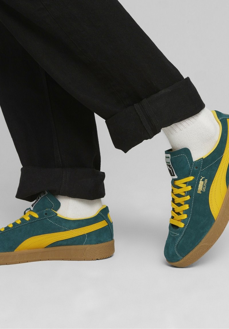 Green and yellow suede sneakers with rubber soles, featuring contrasting stripes and laces, paired with black denim pants and white socks.