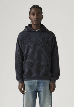 Mann mit Zöpfen, der einen dunkel gemusterten Levi's Hoodie und hellblaue, zerrissene Jeans trägt, steht vor einem einfarbigen Hintergrund.