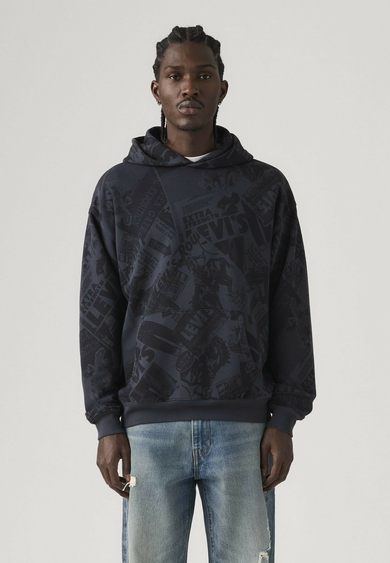 Man with braids wearing dark patterned Levi's hoodie and light blue ripped jeans standing against plain background.