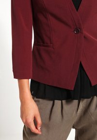 Femme portant un blazer bordeaux ajusté avec un seul bouton, un chemisier noir et un pantalon marron ample, main détendue le long du corps.