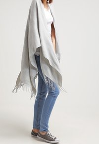 Poncho grigio chiaro con bordi frangiati, realizzato in tessuto morbido. Indossato sopra una maglietta bianca e abbinato a jeans blu e sneakers grigie.