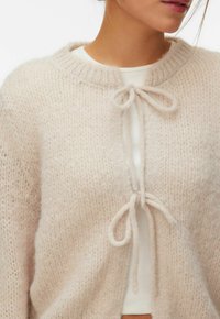 Gros plan sur une personne portant un cardigan beige doux en tricot, noué avec deux nœuds, par-dessus un haut blanc côtelé, montrant le cou et les lèvres.