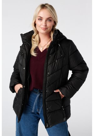 Femme portant une doudoune noire matelassée avec capuche, un pull bordeaux à col en V et un jean bleu, debout les mains dans les poches de la veste.