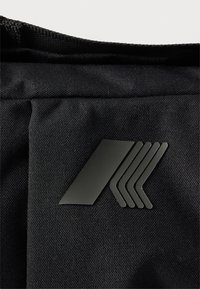 Μαύρο ύφασμα με γυαλιστερό μαύρο λογότυπο που απεικονίζει μία μεγάλη διαγώνια ρίγα και τέσσερις μικρότερες ρίγες σε σχήμα βέλους σε μια τσέπη τσάντας.