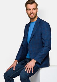 Niebieska marynarka w kratę z klapami, połączona z niebieską koszulą z okrągłym dekoltem i ciemnymi dżinsami. Zauważalny detal guzika na marynarce.