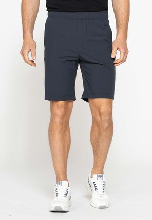 Uomo che indossa pantaloncini blu navy fino al ginocchio e sneakers bianche, in piedi davanti a uno sfondo chiaro e semplice.