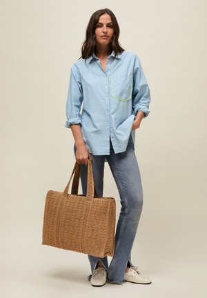 Vrouw staat, draagt een lichtblauw overhemd met knopen, blauwe jeans met voorste splitsingen, witte schoenen, houdt een grote bruine geweven tas vast.