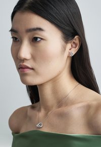 Sudraba kaklarota ar savērptiem apaļiem kuloniem, valkāta virs olīvzaļas plecus atsegjošas topiņas, papildināta ar kniedes auskariem.