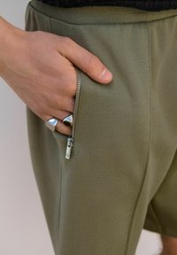Maslinasto zelene kratke hlače od rastezljivog materijala imaju dzep s patentnim zatvaračem i srebrnim hardverom. Ruka je smještena unutar džepa. Glatka tekstura.