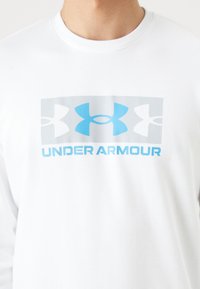 Camisa blanca de manga larga hecha de algodón, con un gran logo de Under Armour en gris y azul en el frontal. Textura suave, cuello redondo.