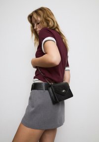 Borsa a tracolla in pelle nera con una catena argentata, forma rettangolare, pattina frontale con un accento metallico. Indossata con un top bordeaux e una gonna grigia.