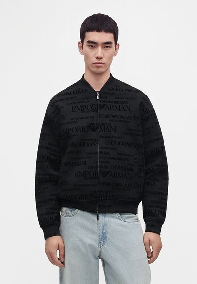 Νέος άνδρας που φοράει μαύρο μπουφάν Emporio Armani με φερμουάρ και λογότυπα σε όλο το ρούχο και ανοιχτόχρωμο μπλε τζιν, στέκεται μπροστά σε απλό λευκό φόντο.