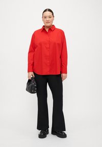 Camicia rossa con bottoni, maniche lunghe, abbinata a pantaloni neri a campana e scarpe nere chunky. Il modello tiene una piccola borsa nera con dettagli di occhielli.