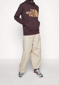 Sudadera con capucha marrón con un gran logo naranja de "THE NORTH FACE", combinada con pantalones cargo beige y zapatillas grises con detalles rojos.