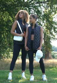 Duas modelos usam coletes acolchoados pretos, leggings pretas e ténis brancos. Uma delas carrega uma pequena bolsa branca; uma coleção branca está na cabeça da segunda.