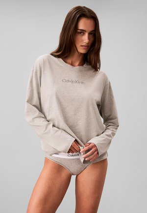 Vrouw draagt een lichtgrijze Calvin Klein top met lange mouwen en bijpassend ondergoed, staand tegen een effen lichtgrijze achtergrond.