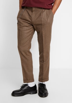 Man draagt bruine geruite op maat gemaakte broek met voorplooien, zwarte leren schoenen, witte sokken en een kastanjebruin overhemd, handen in de zakken.