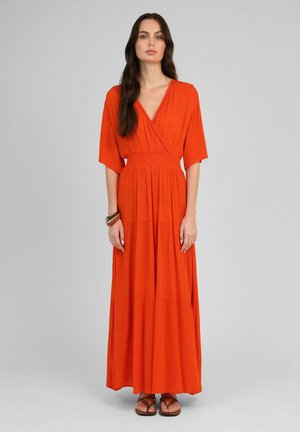 Femme aux longs cheveux foncés portant une robe maxi orange à encolure en V et manches aux coudes, debout devant un fond uni.