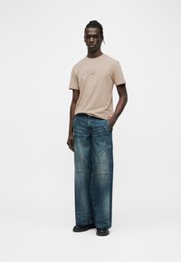 Άνδρας που στέκεται φορώντας μπεζ μπλουζάκι με σχέδια, ξεθωριασμένο μπλε τζιν φαρδιάς γραμμής και μαύρα παπούτσια, σε απλό λευκό φόντο.