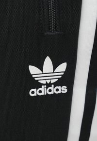 Μαύρα αθλητικά ρούχα με υφή υφάσματος, με λογότυπο Adidas σε λευκό και τρεις λευκές ρίγες κατά μήκος μιας πλευράς. Ορατή λεπτομέρεια φερμουάρ.