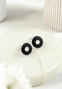 Svarta cirkulära örhängen med en central klar ädelsten, fästa i silverhardware. Slät yta med en glänsande finish.