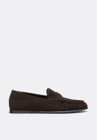 Mocassin pour homme en daim marron foncé avec détails cousus style mocassin et semelle plate noire, présenté de profil sur fond blanc.