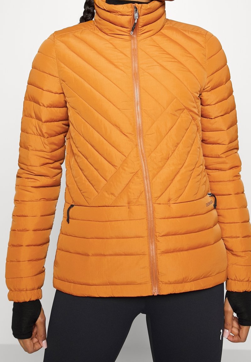Femme portant une veste matelassée orange avec des coutures diagonales et une fermeture éclair frontale, associée à des leggings noirs et des gants sans doigts.