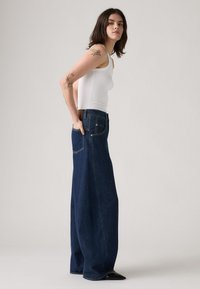 Jeans en denim taille haute et jambes larges de couleur bleu foncé, avec une poche avant, une poche arrière et un accent en cuir marron au niveau de la taille.