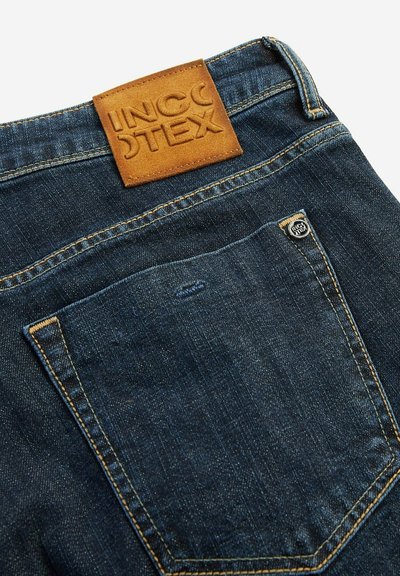Vaqueros de denim azul oscuro con una etiqueta de cintura de cuero marrón texturizado. Presenta un bolsillo trasero con costuras sutiles y una etiqueta de metal.