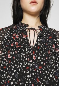 Černá květinová blůza s volánkovým límcem, která má zavazování u krku. Látka je lehká s jemnou, průsvitnou texturou.