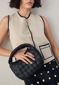 Sac à main en cuir noir tressé avec une poignée tressée, associé à un top beige sans manches avec des boutons et une poche poitrine.